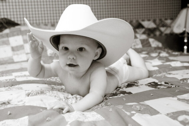 Baby wearing an oversized white cowboy hat lying on a patchwork quilt, black and white