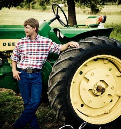 Young man in a plaid shirt posing beside a vintage green John Deere tractor