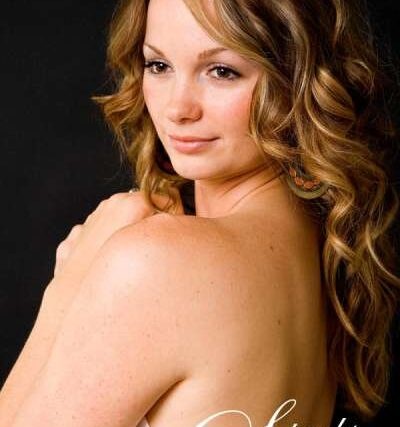 Redheaded woman wrapped in white satin drapery smiling over her bare shoulder