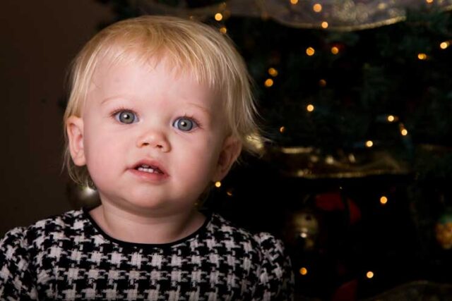 Smiling blond toddler in a houndstooth vest in front of a lit Christmas tree