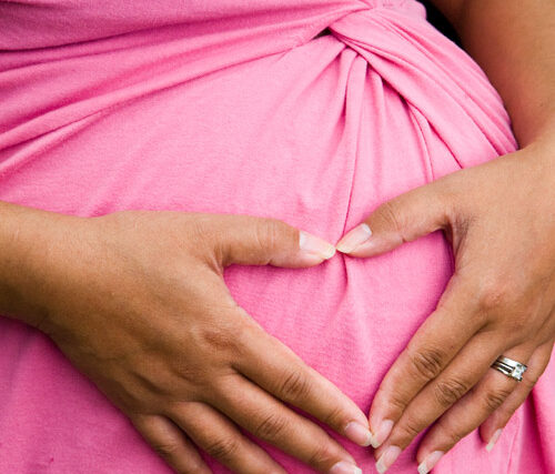 Close-up of a pregnant belly in a pink knot-front dress with manicured hands and a ring