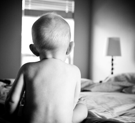 Toddler sitting on a bed gazing at the window in soft light, black and white vignette