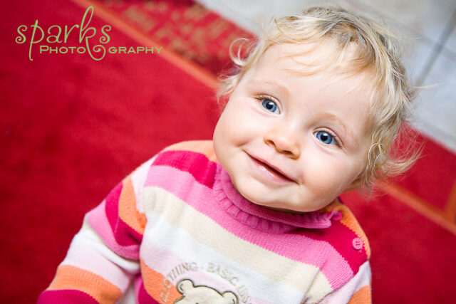Cheerful curly-haired toddler in a pink striped sweater tilting her head on a red carpet