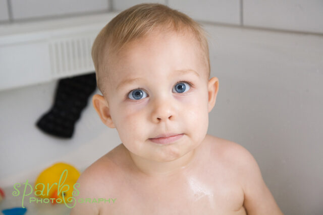 Blue-eyed baby sitting in a bathtub splashing in soapy water, color