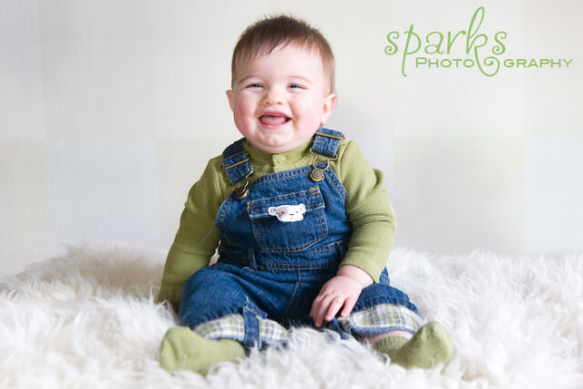 Giggling baby in green sleeves and denim overalls sitting on white sheepskin