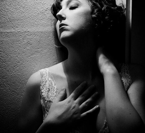 Black and white boudoir portrait of a woman in lace lingerie leaning against a wall with eyes lowered