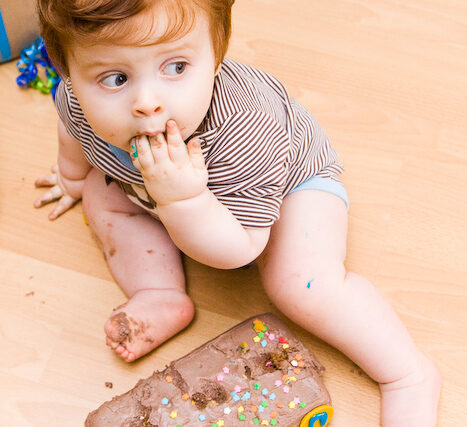 Baby sitting on the floor sucking his fingers in front of a smashed birthday cake