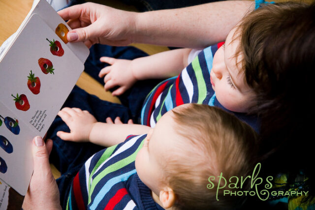 Two toddler boys in striped shirts looking at a strawberry picture book read aloud by their mother