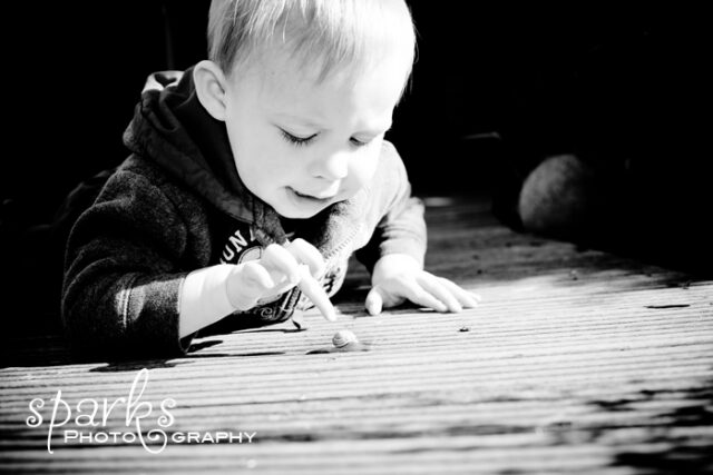 Blond toddler in a hooded sweatshirt kneeling on a porch, watching a garden snail crawl across the boards, selective color