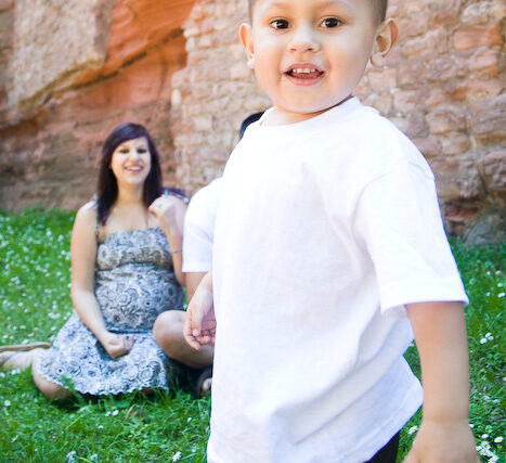 Smiling toddler boy in a white shirt toddling toward the camera while his pregnant mom sits in the grass behind