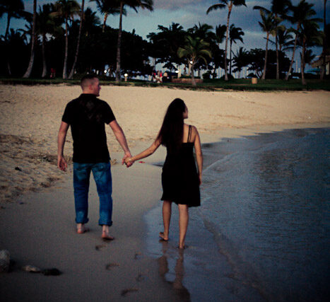 Couple walking hand in hand along a palm-lined beach under evening clouds