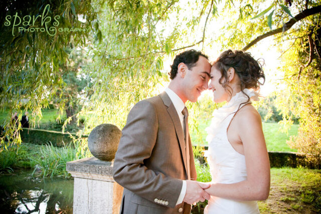 A wedding couple touches foreheads while holding hands beside a stone pillar and pond under a willow tree.