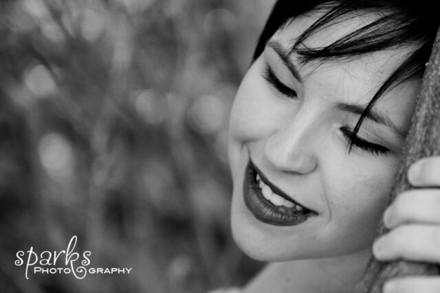 Melissa D smiles with closed eyes as she leans against a tree trunk in a leafy outdoor setting.