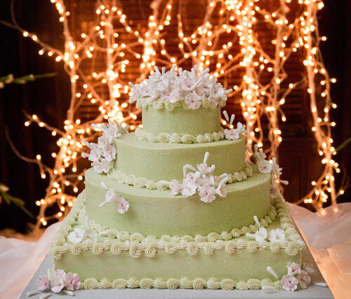 A three-tier light green wedding cake with white flowers sits before glowing string lights.