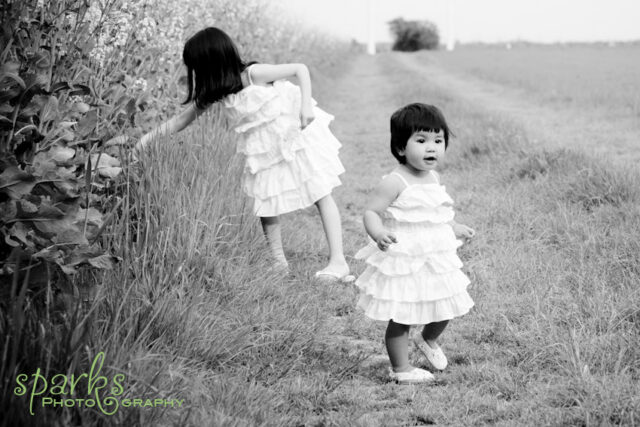 Two young girls in white tiered dresses walk through a grassy field beside tall flowering plants.