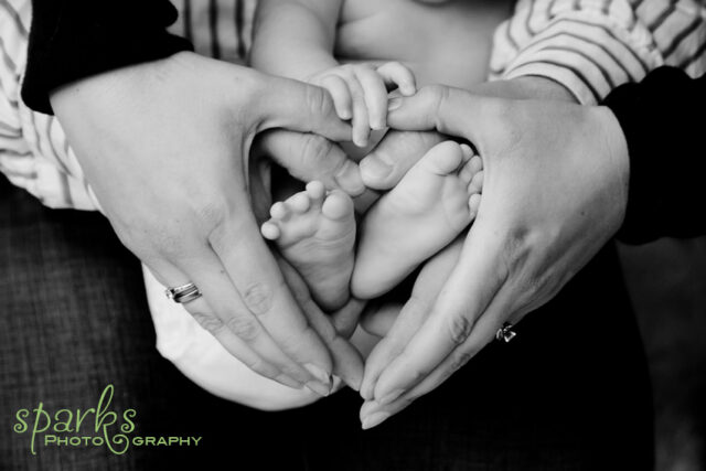 Adult hands form a heart shape around a baby's tiny feet in a tender black-and-white portrait.