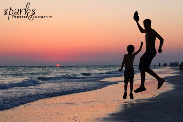 Silhouetted man and child leap on the shoreline as the sun sets over the ocean.