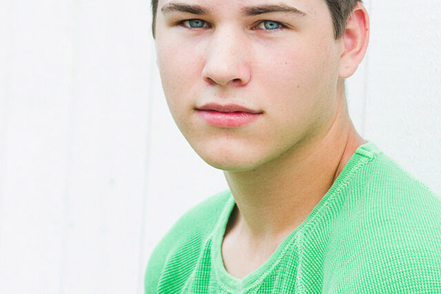 A young man in a green shirt poses for his high school senior portrait against a white paneled wall.