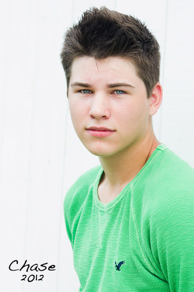 A young man in a green shirt poses for his high school senior portrait against a white paneled wall.