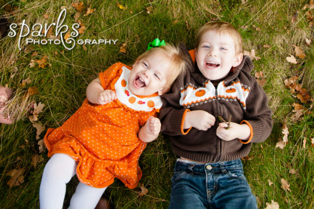 Two laughing children lie side by side on autumn grass scattered with fallen leaves.