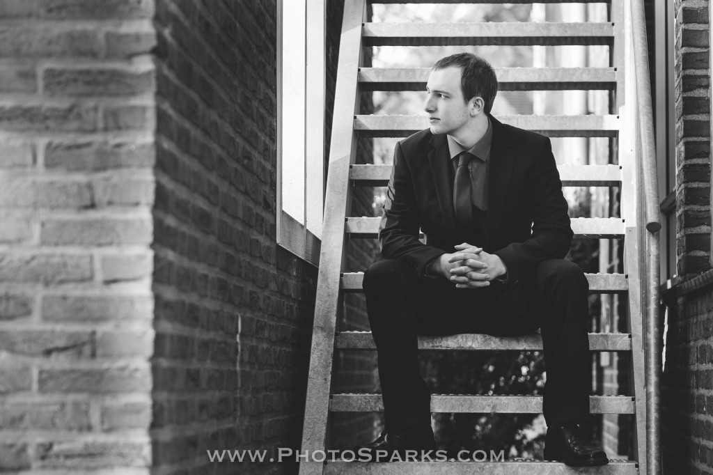 A man in a dark suit sits on a brick staircase with hands clasped in a black-and-white portrait.