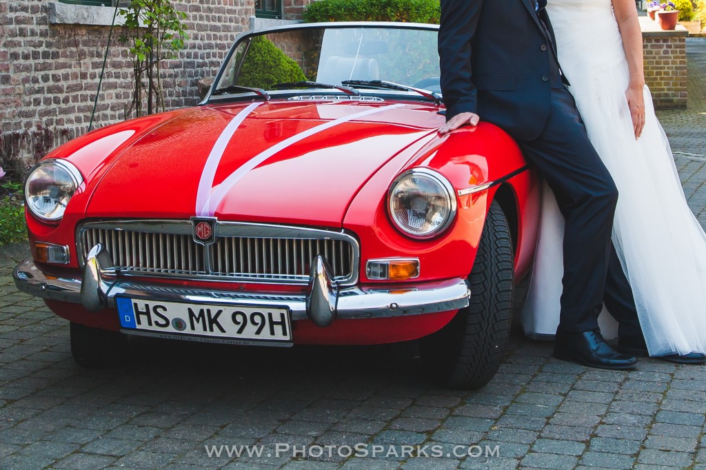 Bride and groom stand beside a red vintage wedding car on a cobblestone courtyard.