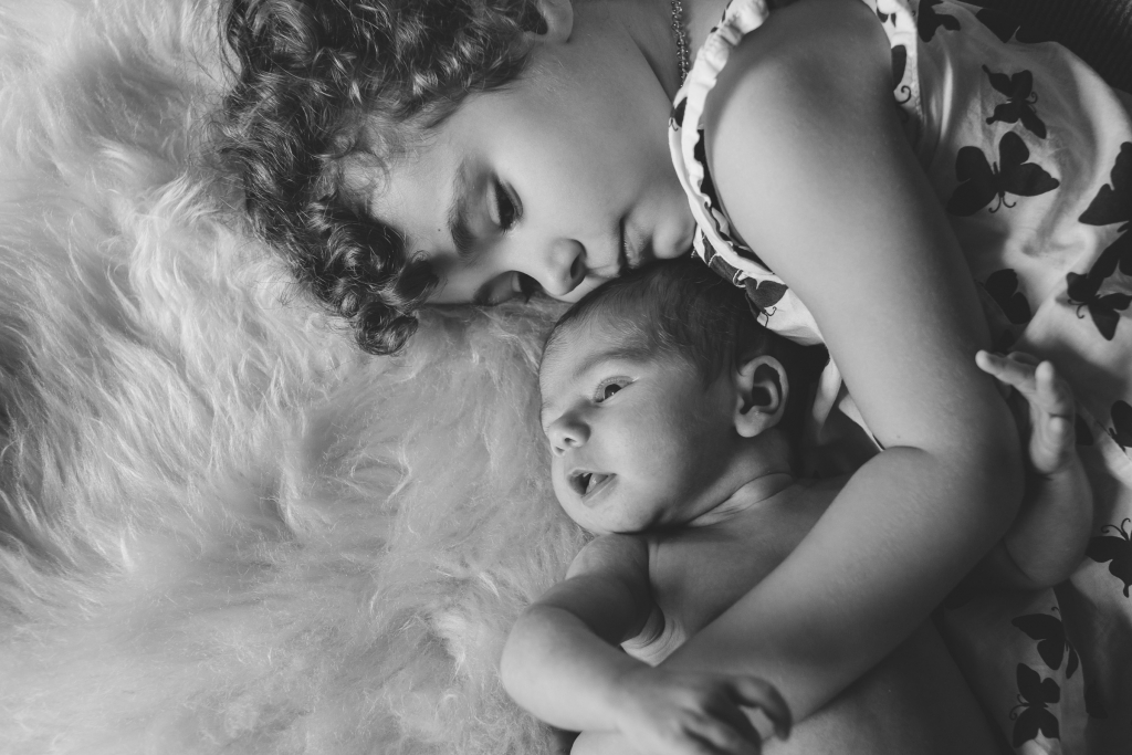 A curly-haired child and a newborn baby rest together on a fluffy white blanket.