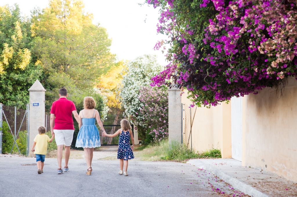 A family of four walks hand in hand down a tree-lined path in Valencia beneath bright purple bougainvillea.