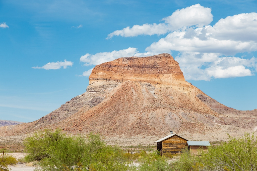 A flat-topped mesa rises above a small wooden cabin and desert shrubs in Big Bend.