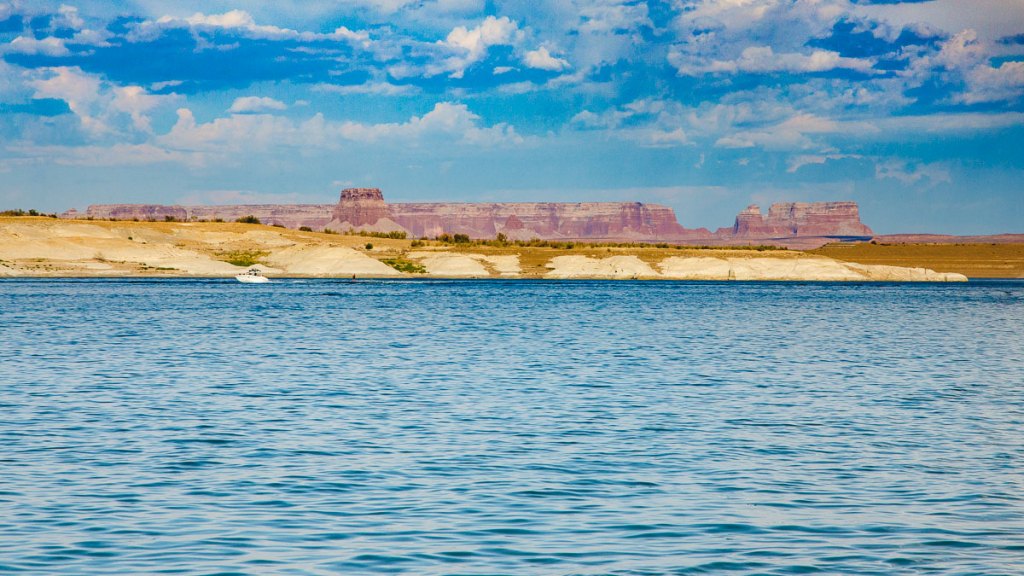 Turquoise lake water in the foreground with a sandy shoreline and distant red canyon mesas at Lake Powell in Page Arizona