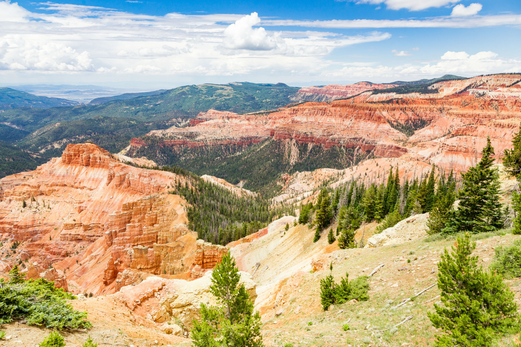 Panorama of Cedar Breaks National Monument with orange eroded slopes descending into a forested canyon under a cloudy blue sky