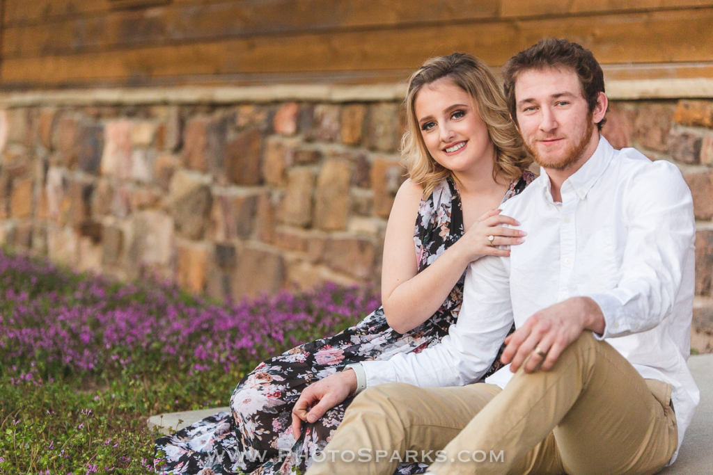 Engaged couple sitting together in front of a stone and wood barn with purple flowers