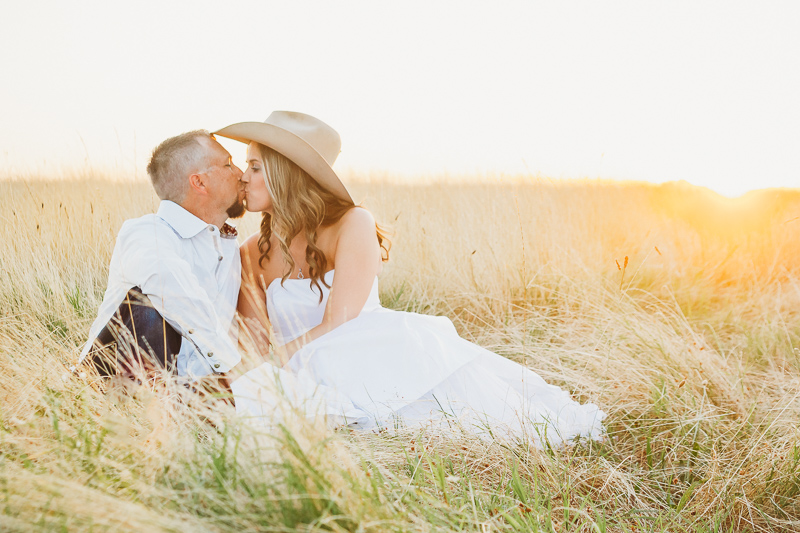 Wedding couple sitting in tall grass at sunset. Bride is wearing a cowboy hat.