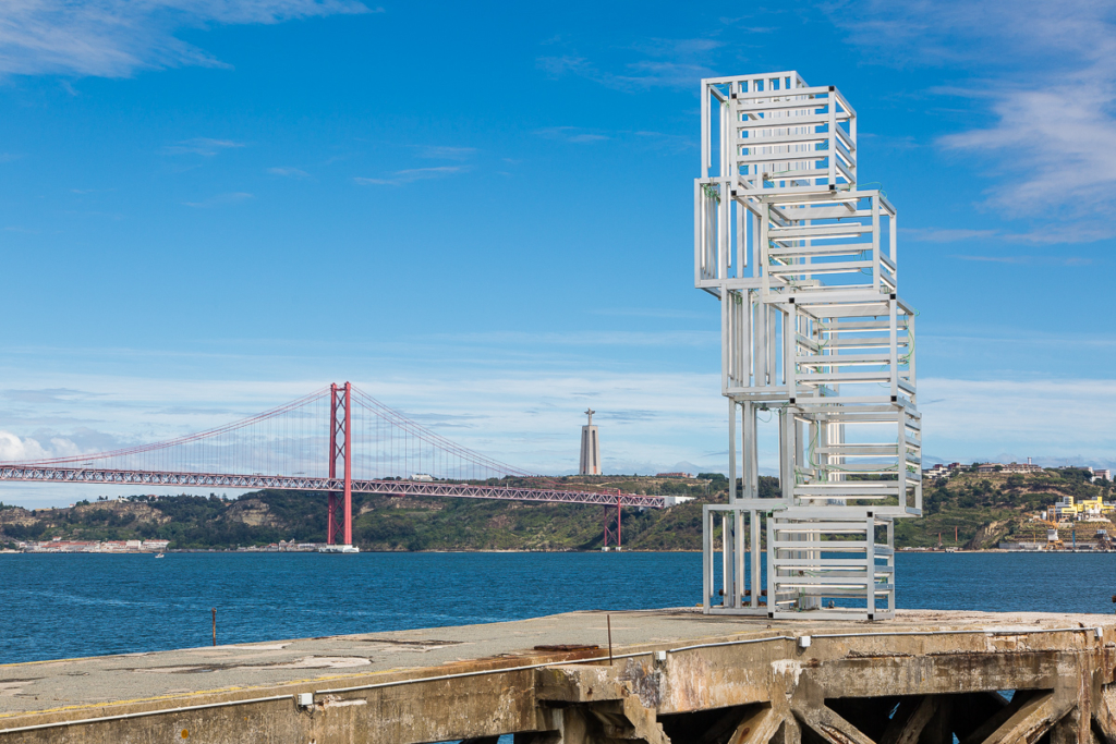 White metal sculpture on a Lisbon pier with the 25 de Abril Bridge in the background
