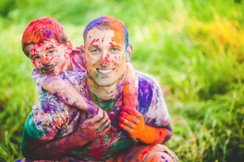 Father carrying his son on his back, both painted rainbow colors after Holi