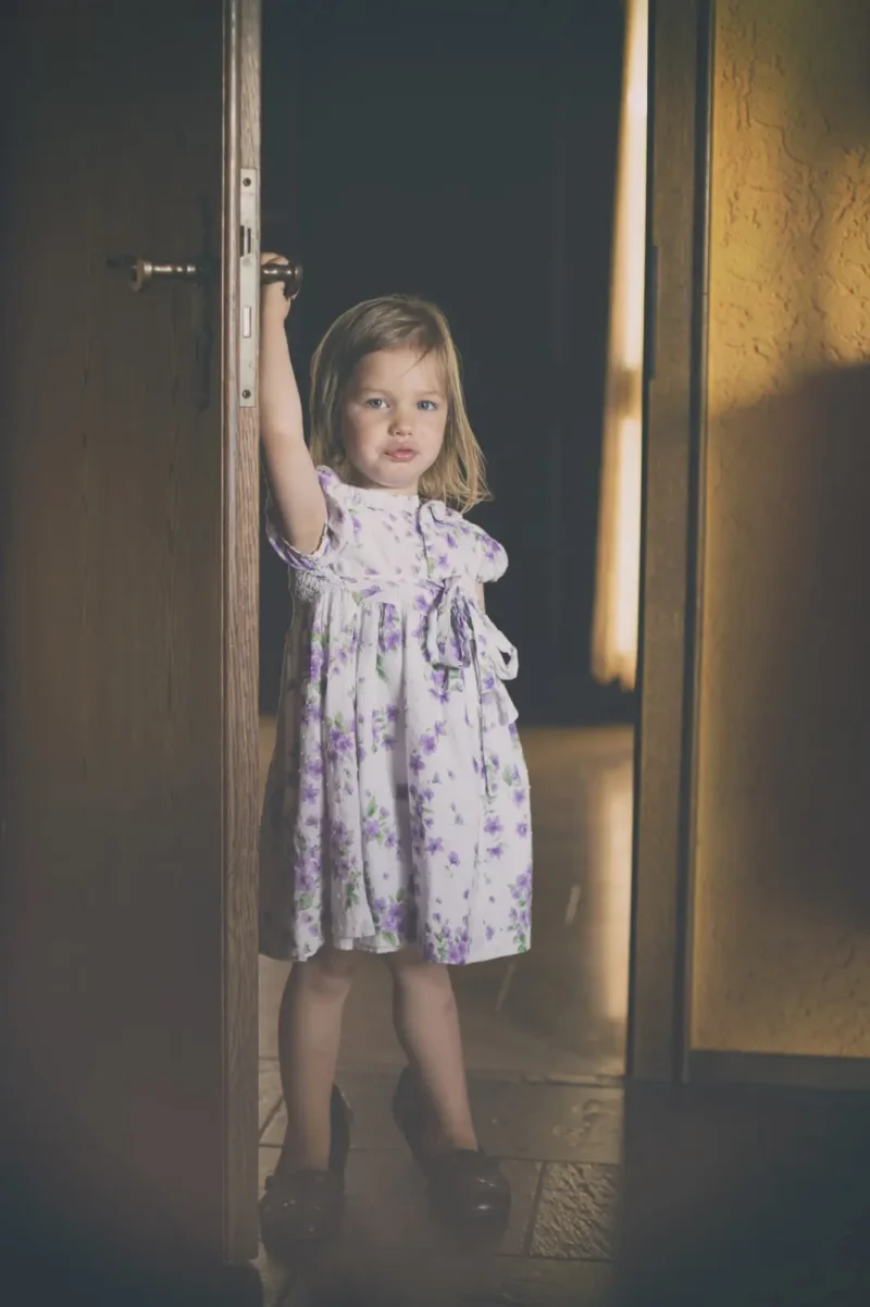 Toddler in a purple nightgown standing in a dim doorway holding the door frame