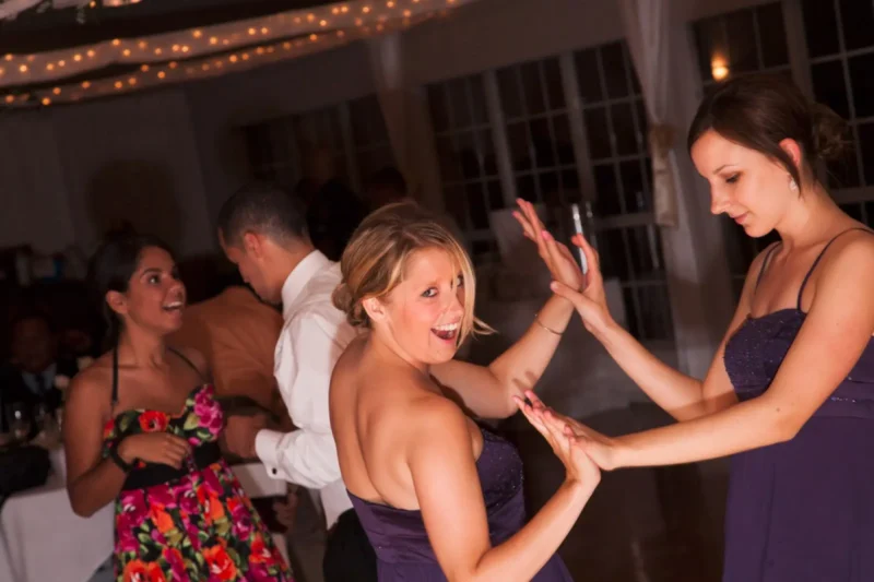 Two women laughing on the dance floor, one touching the other's hair in a string-light reception