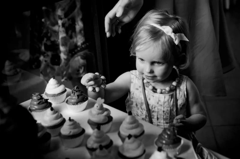 Black-and-white photo of a flower girl examining a tray of wedding cupcakes